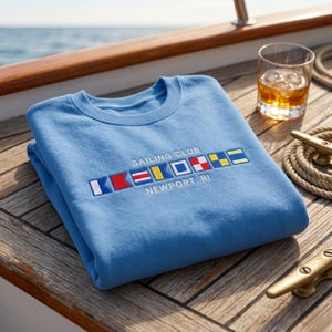 Könnte beinhalten: Hellblaues Sweatshirt mit dem gestickten Text "SAILING CLUB NEWPORT, RI" und bunten nautischen Flaggen. Das Sweatshirt liegt gefaltet auf einem Holzdeck, im Hintergrund ein Glas mit bernsteinfarbener Flüssigkeit und ein Seil.