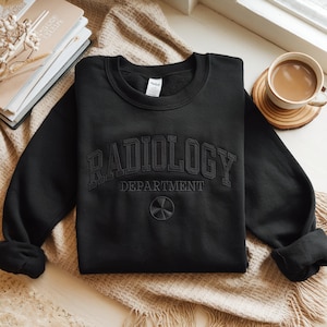 Könnte beinhalten: Schwarzes Sweatshirt mit dem gestickten Schriftzug "RADIOLOGY DEPARTMENT" in fetter Schrift und einem Strahlungssymbol darunter. Das Sweatshirt wird auf einer neutralfarbenen Decke präsentiert.