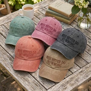 Op de afbeelding: Vijf baseballpetten in verschillende kleuren, waaronder saliegroen, poederroze, koraal, antracietgrijs en beige. Elke pet heeft de tekst "MISS NEVER DO NO WRONG" geborduurd. De petten liggen op een houten tafel.