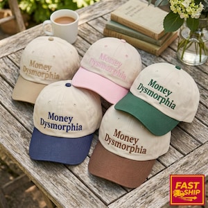 May include: A collection of baseball caps in various colors, including tan, pink, green, brown, and navy blue. Each cap features the embroidered text "Money Dysmorphia" in a contrasting color. The caps are arranged on a wooden surface.