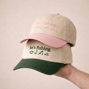 May include: Two beige baseball caps stacked on top of each other. The top cap has a pink bill and embroidery that reads "he's fishing." The bottom cap has a green bill and green embroidery with the same text.
