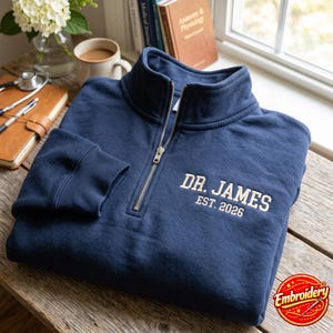 Op de afbeelding: Een marineblauw sweatshirt met een kwartrits, een hoge kraag en een zilverkleurige metalen rits. Op de borst is de witte tekst "DR. JAMES" geborduurd boven de kleinere witte tekst "EST. 2026" in een bloklettertype.