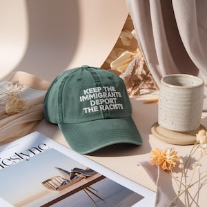 Puede incluir: Gorra de béisbol verde con el texto blanco "KEEP THE IMMIGRANTS DEPORT THE RACISTS". La gorra está colocada cerca de una revista y una taza de cerámica moteada.