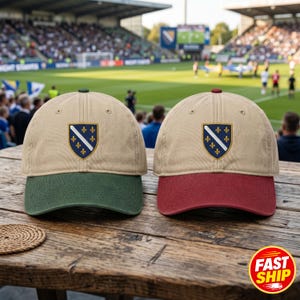 Peut inclure: Deux casquettes de baseball avec des calottes beiges et des écussons brodés présentant une bande diagonale et des fleurs de lys jaunes. L'une a une visière et un bouton vert foncé, l'autre une visière et un bouton rouge foncé, sur une table en bois.
