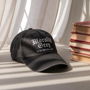 Puede incluir: Gorra de béisbol gris oscuro con la frase "Morally Grey is my favorite color" en letras blancas. La gorra tiene una visera curva y está hecha de un material suave. Se ve una pila de libros en el fondo.