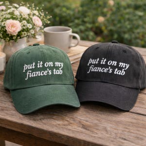 Puede incluir: Dos gorras de béisbol, una verde oscuro y otra negra, sobre una superficie de madera. Ambas gorras tienen la frase bordada "put it on my fiance's tab" en blanco. Un arreglo floral y una taza son visibles.