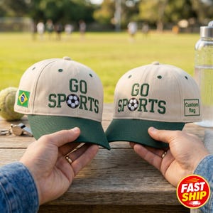 May include: Two beige baseball caps with dark green brims and dark green embroidered text that reads GO SPORTS, featuring a soccer ball graphic in place of the letter O. One cap displays a Brazilian flag patch, while the other has a custom flag label.
