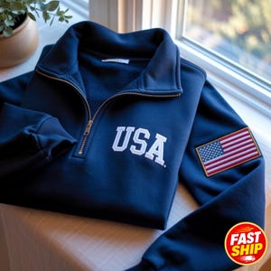 Könnte beinhalten: Ein marineblaues Sweatshirt mit kurzem Reißverschluss, weiß gesticktem USA-Schriftzug auf der Brust und einem rechteckigen Aufnäher der amerikanischen Flagge am linken Ärmel. Das Kleidungsstück hat einen hohen Kragen und gerippte Ärmelbündchen.