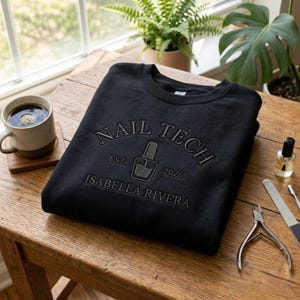 Puede incluir: Sudadera negra con el texto "NAIL TECH" arqueado en la parte superior, con un gráfico de botella de esmalte de uñas y el texto "EST. 2026" debajo. El nombre "ISABELLA RIVERA" está impreso debajo del gráfico. Una taza de café, herramientas de uñas y plantas están sobre la mesa de madera.