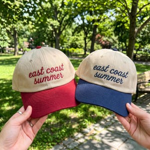 Op de afbeelding: Twee baseballpetten met beige kronen en geborduurde tekst east coast summer. De ene pet heeft een rode klep en rode letters, terwijl de andere pet een marineblauwe klep en marineblauwe letters heeft. Beide petten hebben een knoop bovenop.