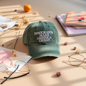 以下が含まれることがあります： 「IMMIGRANTS MAKE AMERICA GREAT」の白い文字がプリントされた、くすんだ緑色のベースボールキャップ。 ライトブラウンの表面に置かれ、メガネ、ノート、ドライフラワーなどの他のアイテムと一緒に配置されています。 キャップは使い古された外観をしています。