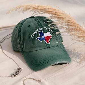 May include: A green baseball cap with the Texas state flag embroidered on the front. The cap features the phrase "Stronger than the Storm" above the flag. The hat is made of a soft material and has a curved brim.