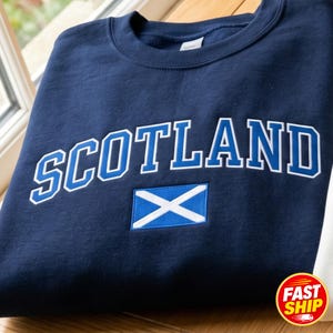 May include: A navy blue crewneck sweatshirt featuring the word SCOTLAND embroidered in bold blue letters with a white outline. Below the text is an embroidered rectangular patch of the Scottish flag, displaying a white saltire on a blue field.