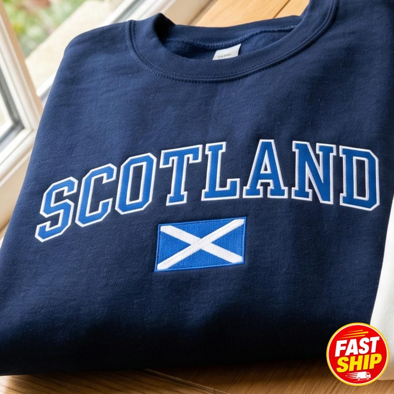 May include: A navy blue crewneck sweatshirt featuring the word SCOTLAND embroidered in bold blue letters with a white outline. Below the text is an embroidered rectangular patch of the Scottish flag, displaying a white saltire on a blue field.
