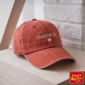 May include: A terracotta baseball cap featuring the embroidered words "chapter 21" and a heart. The cap has a curved peak and a fabric strap. The hat is made of a soft, textured material.