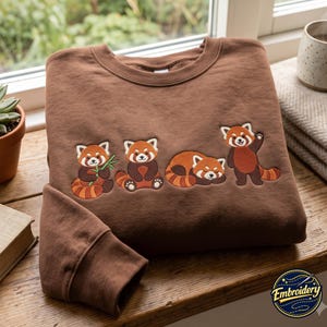 May include: A brown crewneck sweatshirt featuring an embroidered design of four red pandas in various poses. One red panda holds bamboo, another sits upright, one is curled up sleeping, and the fourth stands with one paw raised in a wave.