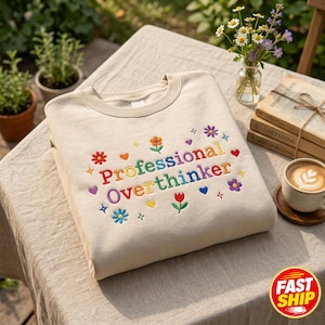 Puede incluir: Una sudadera beige de cuello redondo con el texto bordado "Professional Overthinker" en letras multicolores. El diseño está rodeado de pequeñas flores, corazones y estrellas bordadas en tonos rojo, naranja, amarillo, azul y morado.