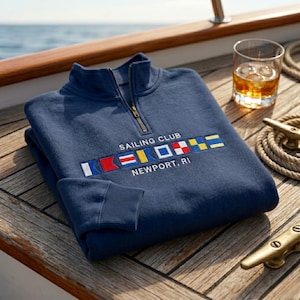 Pode incluir: Um moletom azul marinho Sailing Club Newport, RI com gola com zíper. O moletom apresenta bandeiras de sinalização náuticas bordadas e texto. Um copo de líquido âmbar e uma corda estão no fundo.