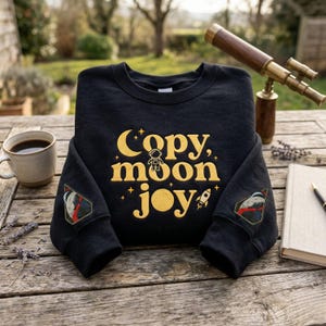 Puede incluir: Sudadera negra con las palabras "Copy, Moon, Joy" en letras doradas, con un astronauta y un cohete. Las mangas tienen parches con un diseño espacial. Una taza de café, un telescopio y un libro están sobre una mesa de madera.
