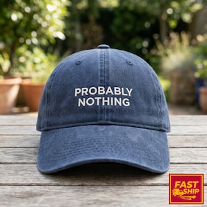 May include: A navy blue baseball cap with the text "PROBABLY NOTHING" in white. The cap is made of a soft material and has a curved brim. The background is a wooden surface with plants.