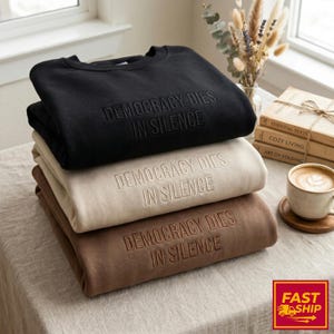 Op de afbeelding: Een stapel van drie sweatshirts in zwart, beige en bruin. Elk sweatshirt heeft de geborduurde tekst "DEMOCRACY DIES IN SILENCE". De sweatshirts zijn netjes opgevouwen en op elkaar gestapeld.