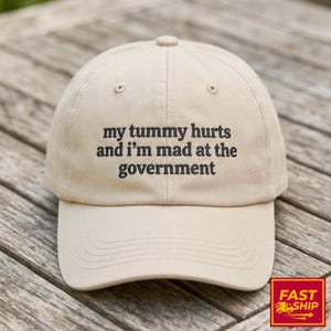 May include: A beige baseball cap with the text "my tummy hurts and I'm mad at the government" in black. The cap has a curved brim and a button on top. The background is a wooden surface.