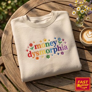 May include: A beige sweatshirt embroidered with the words "money dysmorphia" in a rainbow of colors, surrounded by floral designs. The sweatshirt is folded on a wooden table, with a small vase of flowers and a cup of coffee nearby.