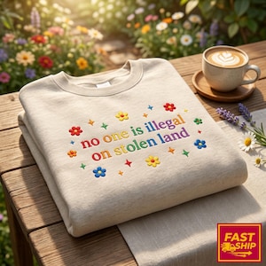 May include: A beige sweatshirt with the embroidered text "no one is illegal on stolen land" in rainbow colours, surrounded by colourful flower and star designs. The sweatshirt is folded on a wooden table.