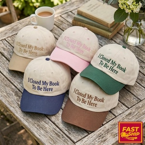 Op de afbeelding: Vijf baseballpetten met gebroken witte kronen en contrasterende kleppen in beige, roze, donkergroen, marineblauw en bruin. Elke pet is voorzien van de geborduurde tekst I Closed My Book To Be Here in de kleur van de bijbehorende klep.