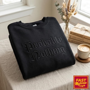 May include: Black crewneck sweatshirt with the embroidered phrase "Probably Nothing" in a gothic font. The sweatshirt is folded on a neutral-colored surface, with a coffee cup and books in the background.