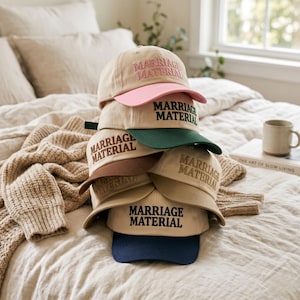 May include: A stack of baseball caps in various colors, including tan, pink, green, brown, and navy blue. Each cap features the embroidered text "MARRIAGE MATERIAL" in a contrasting color. The caps are stacked on a bed with a neutral-toned blanket.