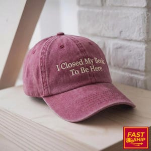 May include: A maroon baseball cap with the embroidered text "I Closed My Book To Be Here". The cap has a curved brim and a button on top. The hat is made of a soft, textured material.