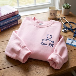 Könnte beinhalten: Hellrosa Sweatshirt mit blauem Stethoskop-Design und dem gestickten Namen "Lisa, RN". Eine Tasse mit "RN", ein Stethoskop und Bücher zum Thema Krankenpflege sind ebenfalls zu sehen.