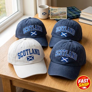 Könnte beinhalten: Drei marineblaue Baseballkappen und eine weiße Baseballkappe, jeweils mit dem Wort SCOTLAND in blauen Buchstaben über einer kleinen gestickten schottischen Flagge. Die Kappen sind auf einer Holzoberfläche neben einer Tasse angeordnet.