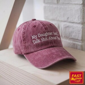 May include: A maroon baseball cap with the text "My Daughter And I Talk Shit About You" in white. The cap is sitting on a wooden surface. The background includes a white brick wall.