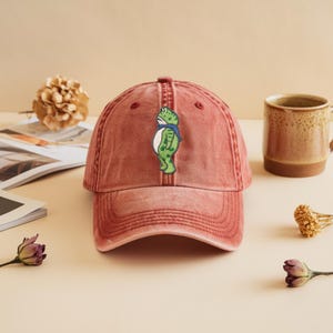 May include: A distressed red baseball cap with a curved peak. The cap has an embroidered green frog wearing a blue scarf. The cap is on a light surface, with a coffee mug and dried flowers in the background.