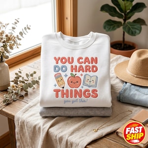 May include: A white crewneck sweatshirt featuring a graphic with the text "YOU CAN DO HARD THINGS" in red and blue, accompanied by illustrations of a pencil, a smiling apple, and an open book, with the smaller text "you got this!" below.