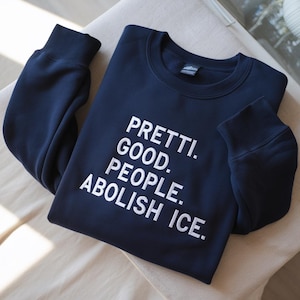 May include: A navy blue sweatshirt with the white text: "PRETTI. GOOD. PEOPLE. ABOLISH ICE." The garment is neatly folded on a light-coloured surface. The sleeves are visible, and the fabric appears soft and comfortable.