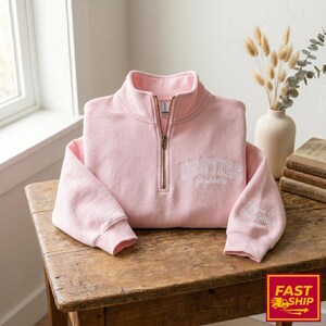 Può includere: Felpa rosa chiaro con zip corta sul collo. La scritta "BESTIES forever" è ricamata sul petto, e sulla manica ci sono i nomi "Olivia & Charlotte". La felpa è appoggiata su un tavolo di legno. Sullo sfondo un vaso e dei libri.