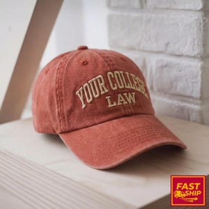 May include: A brick-red baseball cap with the text "YOUR COLLEGE LAW" embroidered in a cream-colored font. The cap is sitting on a wooden surface. The hat has a curved brim and a button on top.