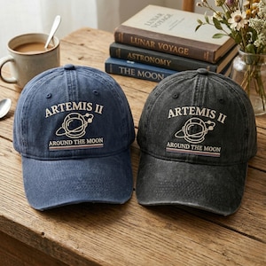 May include: Two baseball caps, one navy blue and one charcoal gray, sit on a wooden surface. Both caps feature the text "ARTEMIS II" above a graphic of a space helmet and the words "AROUND THE MOON". The caps are next to books and a cup of coffee.
