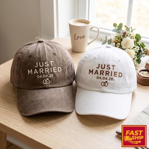 May include: Two baseball caps, one brown and one white, are displayed. Each cap features the embroidered text "JUST MARRIED", the date "04.04.26", and a ring symbol. A mug with the word "Love" is visible in the background.