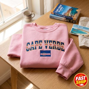 May include: A light pink crewneck sweatshirt featuring the text CAPE VERDE embroidered in a navy blue font with a red and white outline. Below the text is a rectangular patch displaying the national flag of Cape Verde with blue, white, and red stripes and a circle of yellow stars.