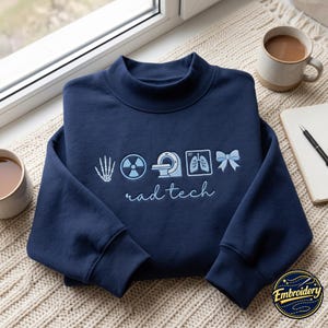 Peut inclure: Un sweat-shirt bleu marine à manches longues avec des broderies bleu clair représentant un squelette de main, un symbole de radiation, un appareil d'IRM, une radiographie pulmonaire et un nœud. Le texte rad tech est brodé en écriture cursive.