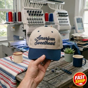 Puede incluir: Una gorra de béisbol beige con visera azul oscuro y el texto American Sweetheart bordado en letra cursiva azul oscuro en el panel frontal. La gorra presenta una corona estructurada con ojales azul oscuro y un botón en la parte superior.