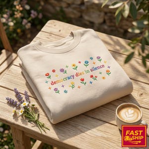 Op de afbeelding: Beige sweatshirt met de tekst "democracy dies in silence" in regenboogkleuren geborduurd, omringd door kleurrijke bloemmotieven. Het sweatshirt ligt op een houten tafel, met een kop koffie en bloemen.