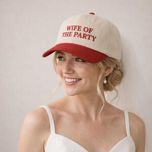 May include: A cream-colored baseball cap with a red brim and the text WIFE OF THE PARTY embroidered in red on the front. The cap features a structured crown, a button on top, and a sheer white veil attached to the back of the headwear.