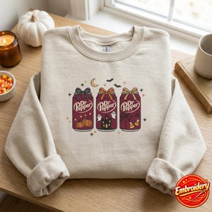Puede incluir: Una sudadera de cuello redondo en color beige con un gráfico de tres latas de refresco Dr. Pepper de color granate. Cada lata tiene un lazo y está decorada con motivos de Halloween como calabazas, una casa encantada, fantasmas y telarañas.