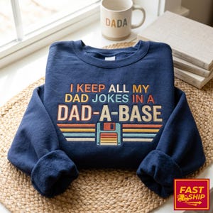 Könnte beinhalten: Marineblaues Sweatshirt mit dem Text "I KEEP ALL MY DAD JOKES IN A DAD-A-BASE" in Orange, Gelb und Weiß. Das Design beinhaltet eine Retro-Diskettengrafik. Eine beige Tasse mit "DADA" ist im Hintergrund zu sehen.
