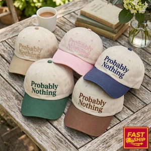 May include: A collection of baseball caps in various colors, including tan, pink, green, brown, and navy blue. Each cap features the embroidered text "Probably Nothing" on the front. The caps are arranged on a wooden surface.